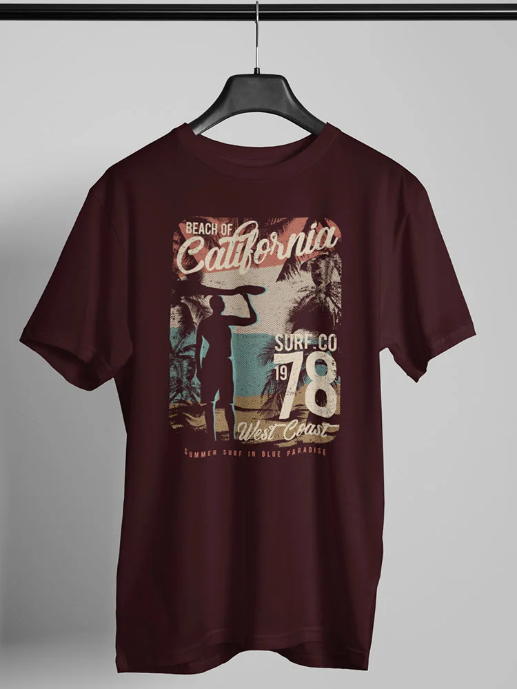 A maroon t-shirt with a vintage California beach design hangs on a hanger, featuring palm trees, a surfer's silhouette, and retro typography.