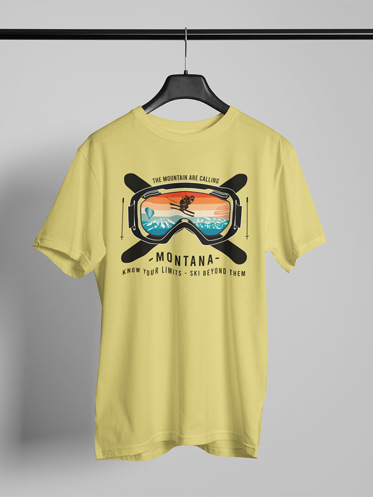 A yellow T-shirt with a ski goggle graphic hangs on a hanger. Text reads "The Mountains Are Calling," "Montana," and "Know Your Limits - Ski Beyond Them."
