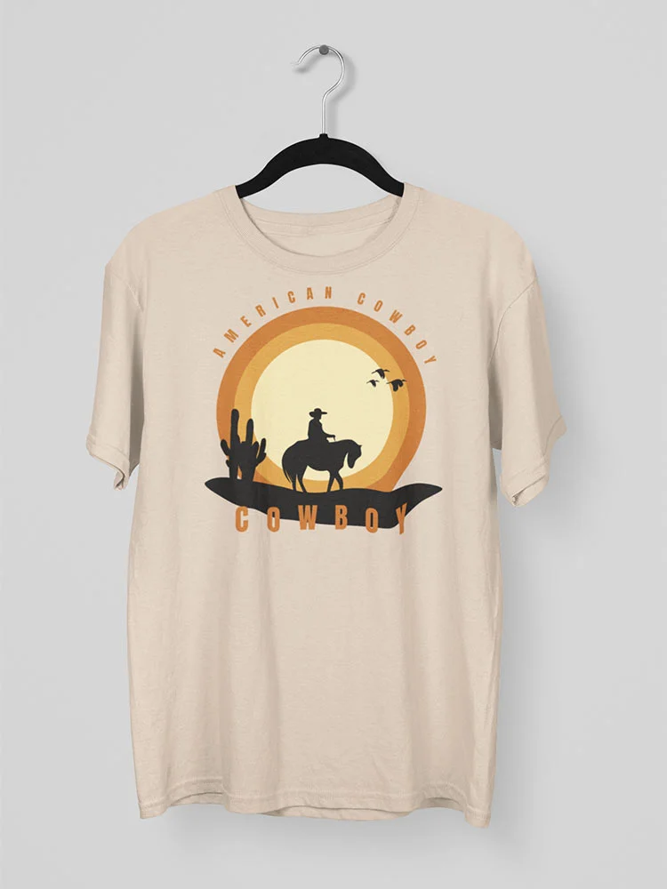 A beige t-shirt with a Western-themed print, featuring a silhouette of a cowboy on horseback, cacti, and birds, against an orange, circular backdrop.