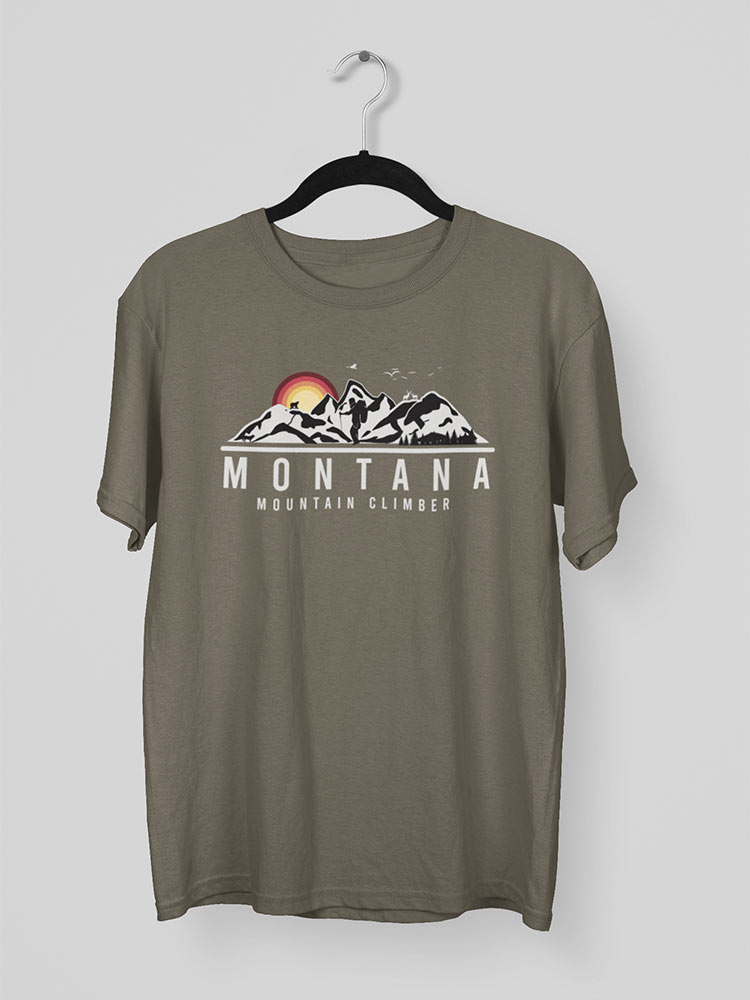 A green t-shirt with a "Montana Mountain Climber" graphic featuring snowy mountains and sunrise hangs on a hanger against a plain background.