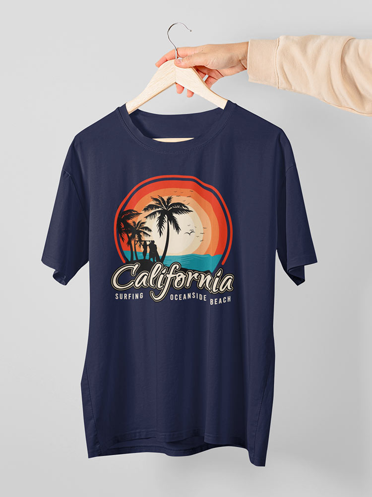 A person is holding a navy blue t-shirt with a California and surfing theme, featuring palm trees, a sunset design, and the words "Oceanside Beach."