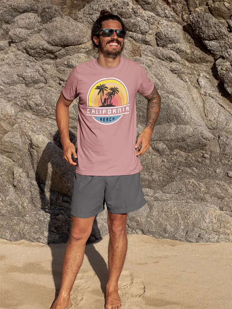 A smiling person wearing sunglasses, a t-shirt, shorts, and no shoes stands on a sandy beach against a rocky background in bright sunlight.