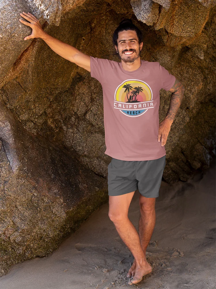 A smiling person is standing by a rock wall at the beach, wearing a brown t-shirt with a "California Beach" logo and grey shorts.