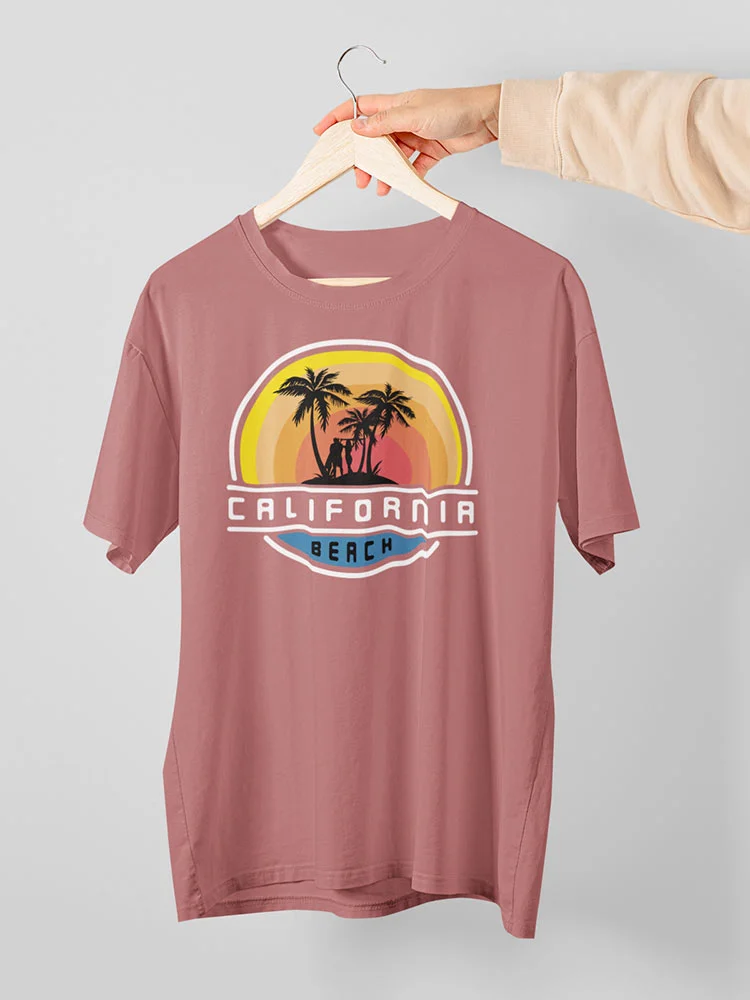 A person's hand holding a dusty rose-colored t-shirt with a "California Beach" print featuring palm trees and sunset on a hanger against a white background.
