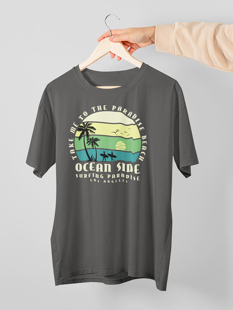 A person is holding a grey t-shirt with a beach-themed graphic design on a wooden hanger against a white background.
