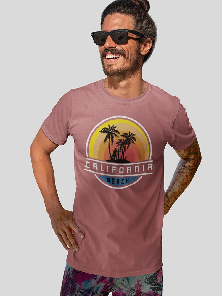 A person wearing sunglasses and a California beach-themed t-shirt is smiling. They have tattoos on their arms and a casual hairstyle.