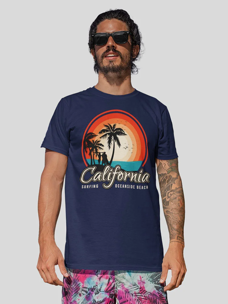 A person with sunglasses and a beard wears a blue t-shirt with a California beach design, standing against a light background, with visible tattoos.