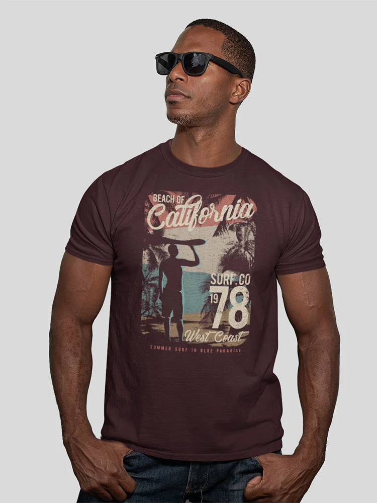 A person stands wearing sunglasses and a brown t-shirt with a vintage California surf-themed graphic, looking confidently to the side.