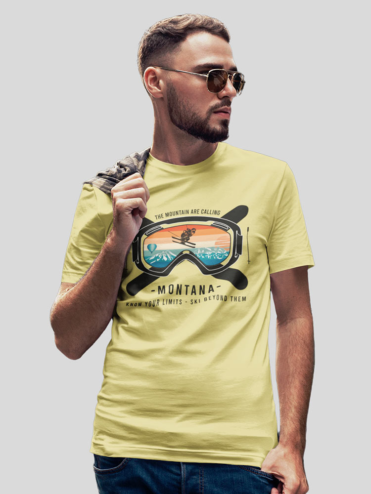 A person posing in a light yellow T-shirt with a graphic of ski goggles and mountains, wearing sunglasses, against a grey background.