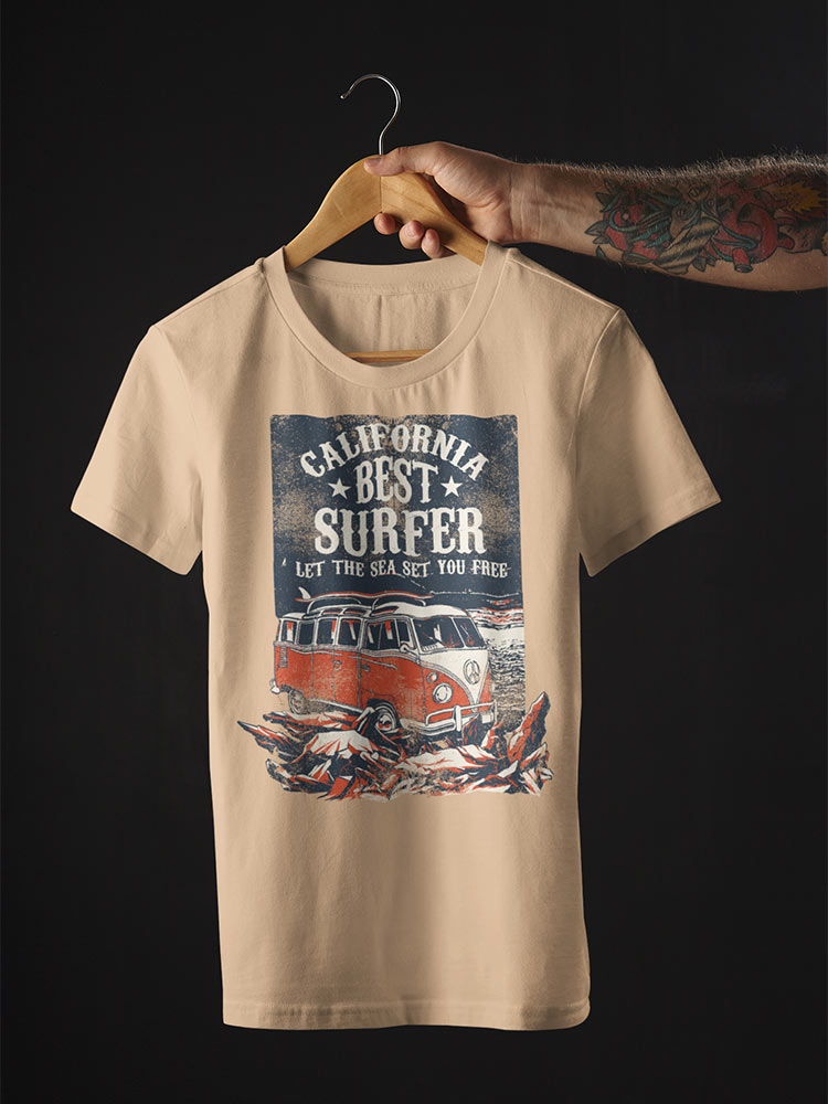 A person's tattooed arm holds a wooden hanger with a beige T-shirt featuring a "California Best Surfer" print with a vintage van and waves.