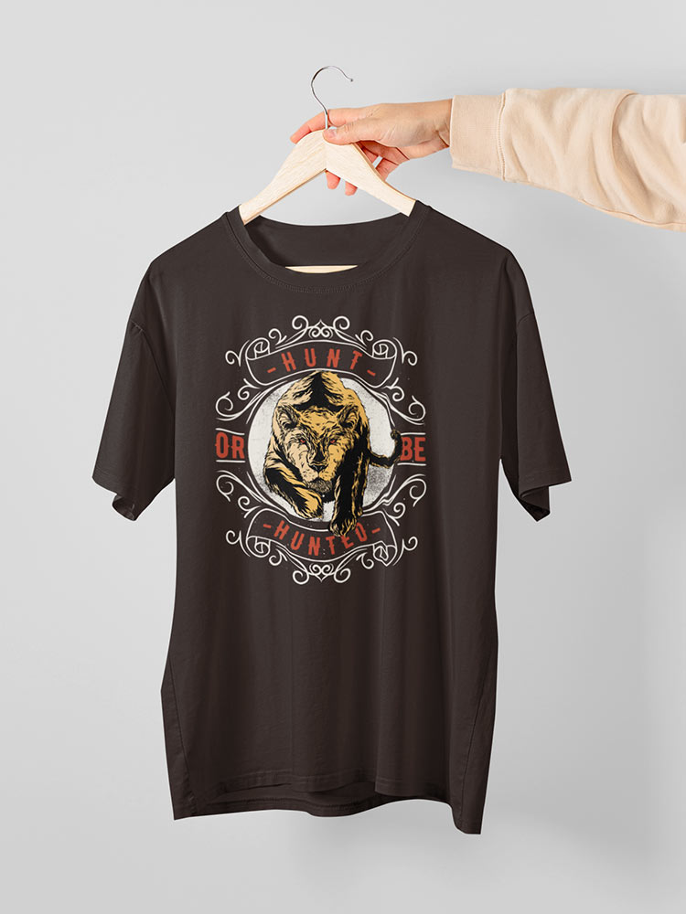 A person is holding a dark t-shirt with a tiger graphic and the words "Hunt or Be Hunted" on a hanger against a light gray background.