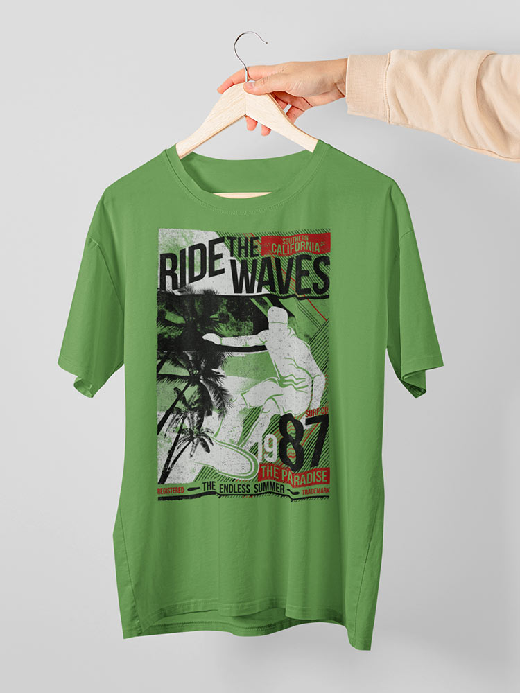 A green T-shirt with a surfing-themed print is displayed on a wooden hanger held by a person's hand against a white background.