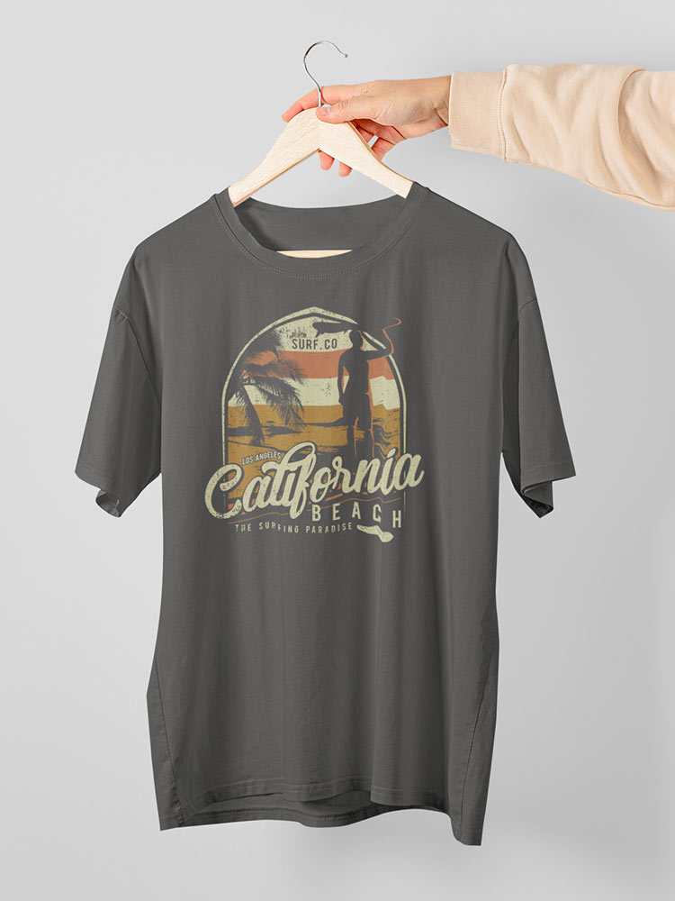 A person's hand is holding a gray t-shirt featuring a "California Beach" graphic with palm trees and a surfboard on a wooden hanger against a white background.
