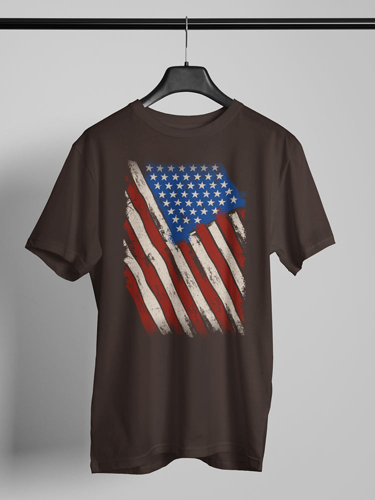 A brown T-shirt with a distressed American flag graphic hangs on a hanger against a grey background, symbolizing patriotism with a vintage style.