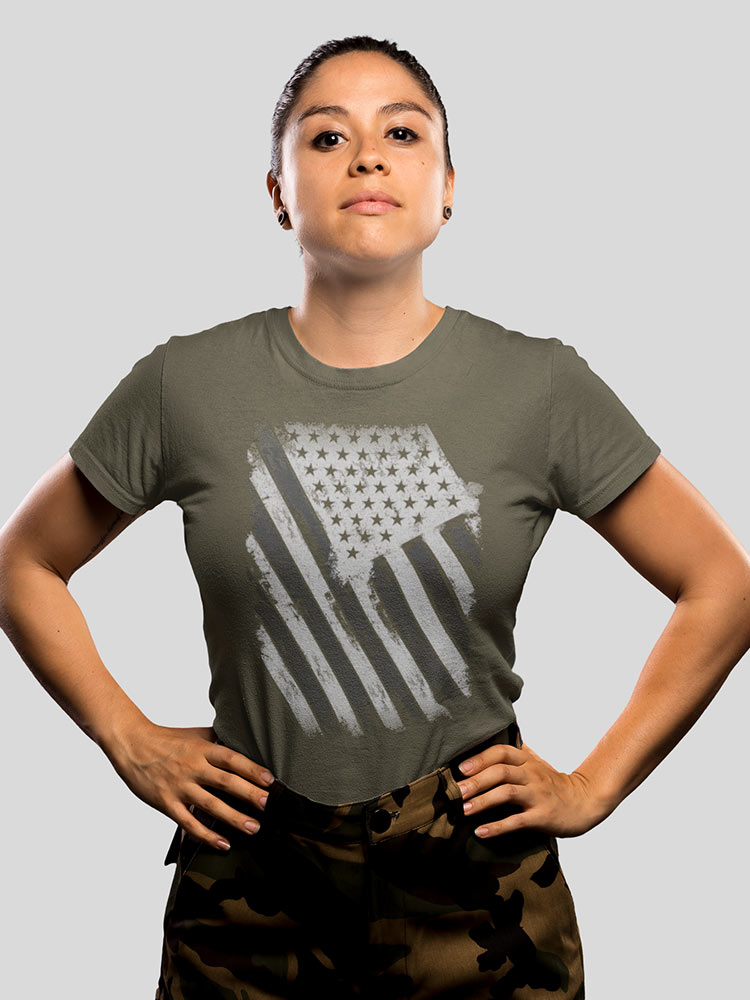 A person stands confidently with hands on hips, wearing a green t-shirt with an American flag print and camouflage pants, against a grey background.
