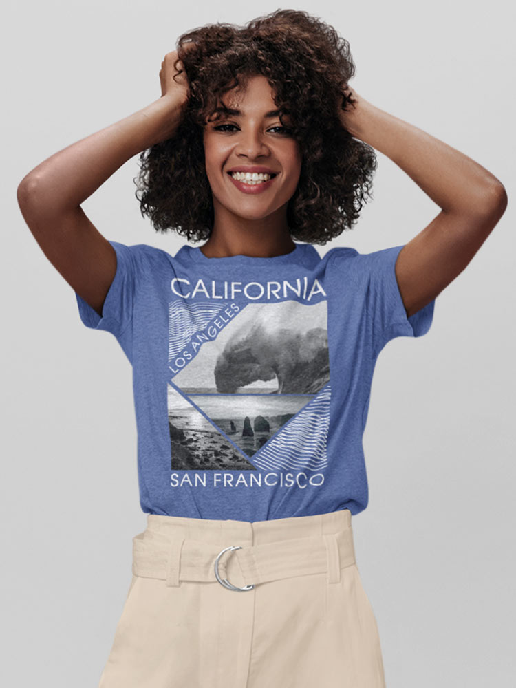 A smiling person with curly hair, wearing a blue "California" T-shirt, stands against a gray background, hands playfully in their hair.