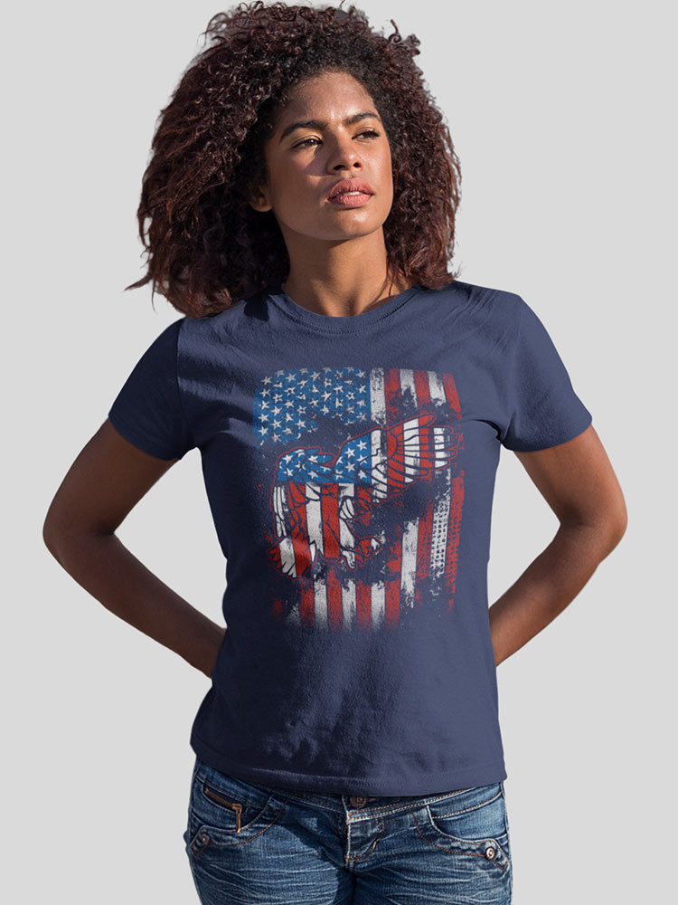 A person with curly hair wearing a blue t-shirt with an American flag and liberty statue print, paired with denim jeans, stands against a light background.