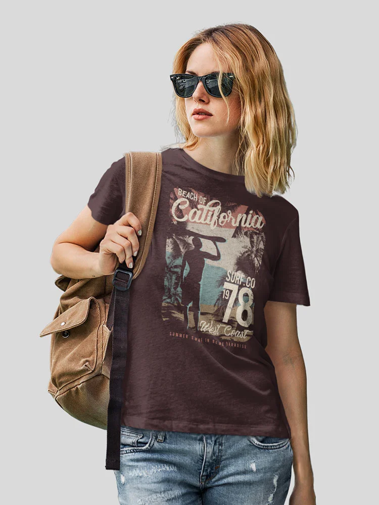 A person with blond hair, wearing sunglasses, a graphic t-shirt, and denim, casually holding a shoulder bag, posed against a grey background.