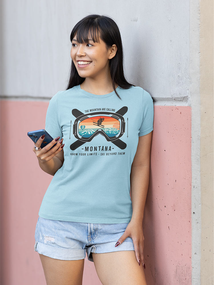 A person leaning against a wall, smiling, holding a smartphone, wearing a T-shirt with a Montana skiing graphic, and denim shorts.