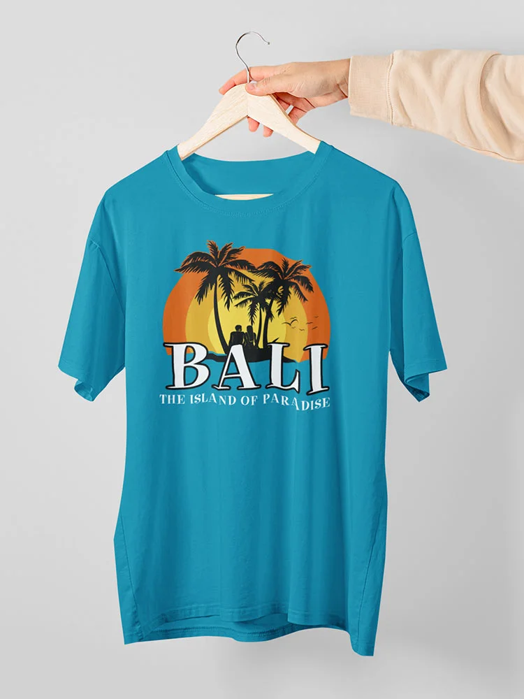 A person is holding a blue t-shirt with a "Bali - The Island of Paradise" design featuring palm trees, sun, and beach on a clothes hanger.