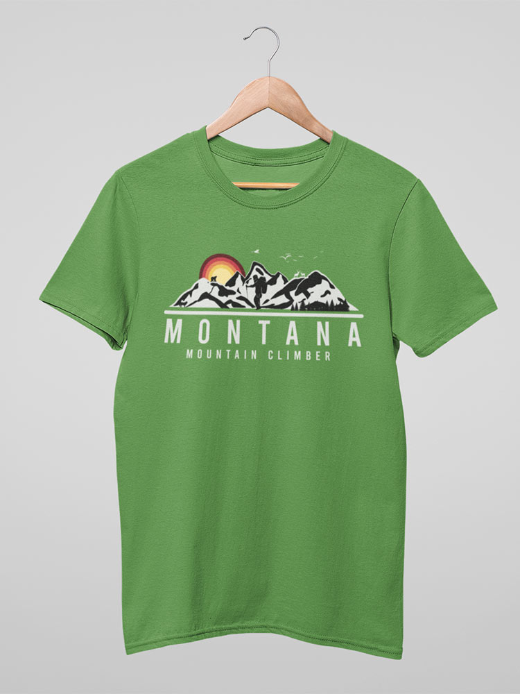 A green t-shirt with a "Montana Mountain Climber" graphic is hanging on a wooden hanger against a white background. The shirt features mountain imagery.