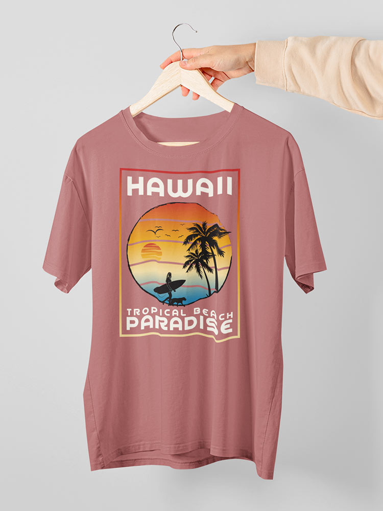 A person is holding up a pink T-shirt with a "Hawaii Tropical Beach Paradise" print showcasing palm trees, a sunset, and a surfer silhouette on a hanger.