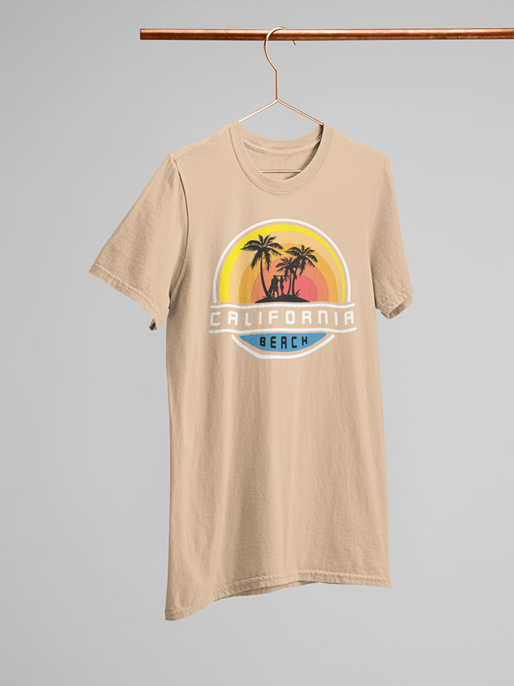 A beige t-shirt with a California Beach theme hangs on a copper hanger against a gray background, featuring palm trees and sunset graphics.