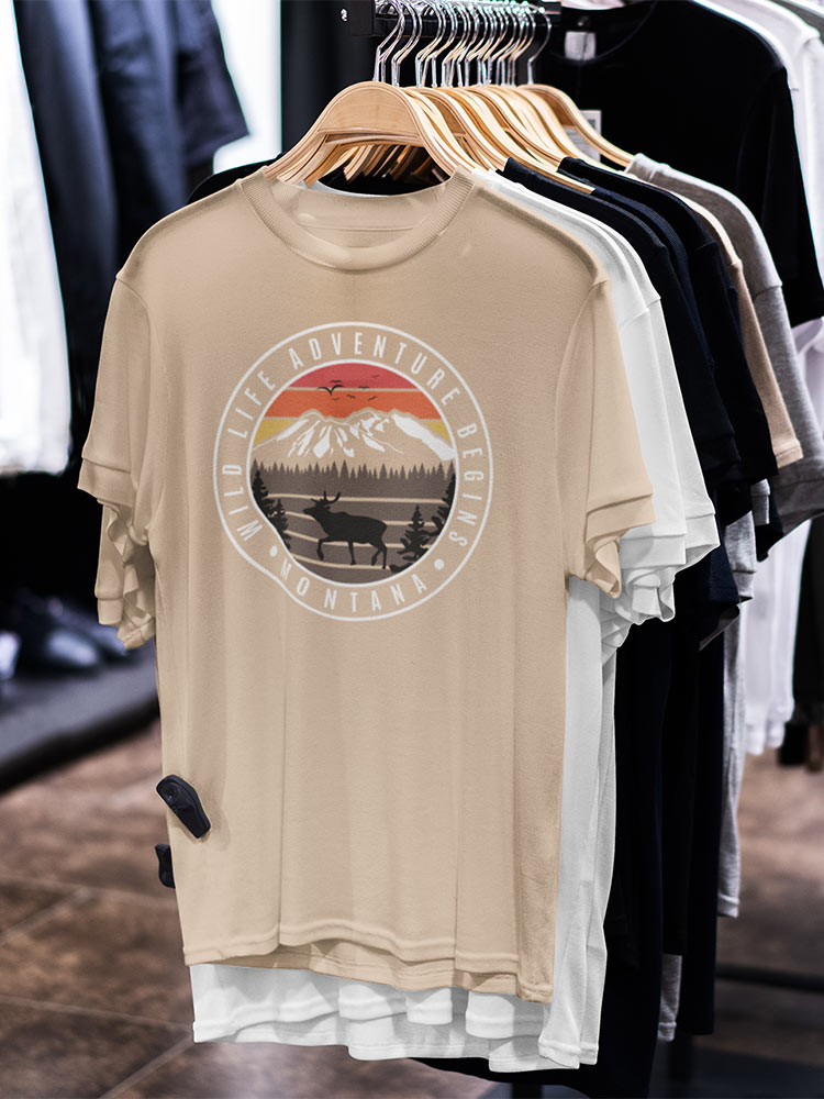 A beige T-shirt with a mountain and wildlife design hangs in a store among other shirts. The text reads "Life Adventure Begins - Montana".