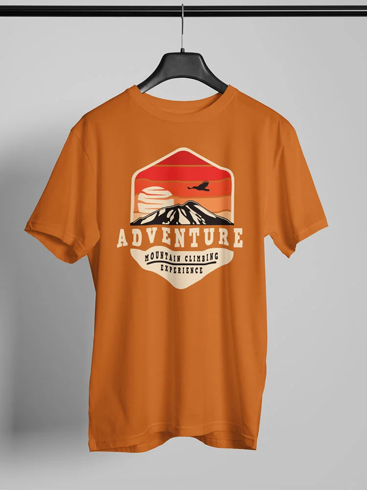An orange t-shirt with an "Adventure - Mountain Climbing Experience" design hangs on a hanger against a plain background, featuring a silhouette of a mountain and bird.