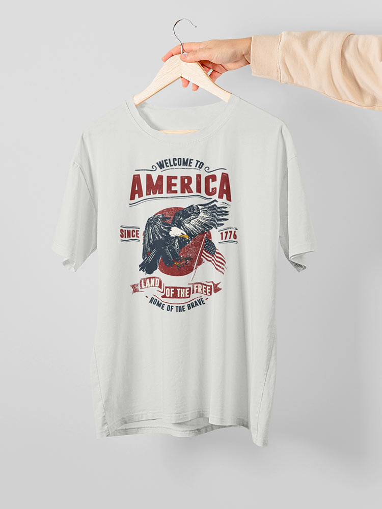 A person holds a white T-shirt with a patriotic American eagle print, featuring the phrases "Welcome to America" and "Land of the Free."