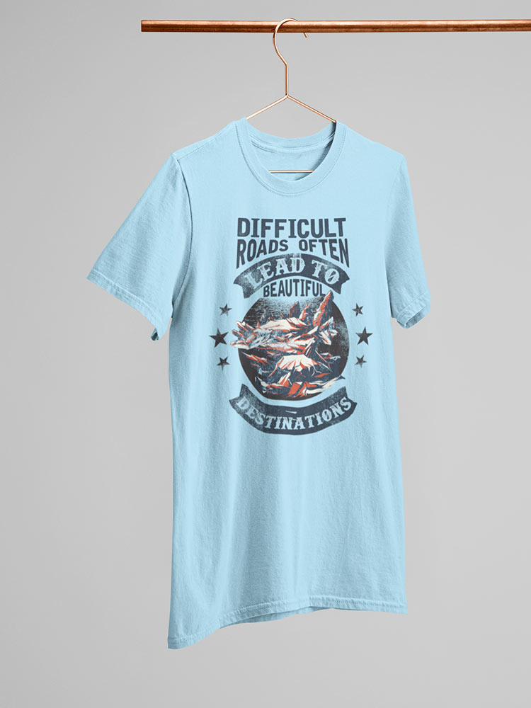 A light blue t-shirt hangs on a wooden hanger against a neutral background, featuring an inspirational quote about overcoming difficult paths, accompanied by stars and a mountain graphic.