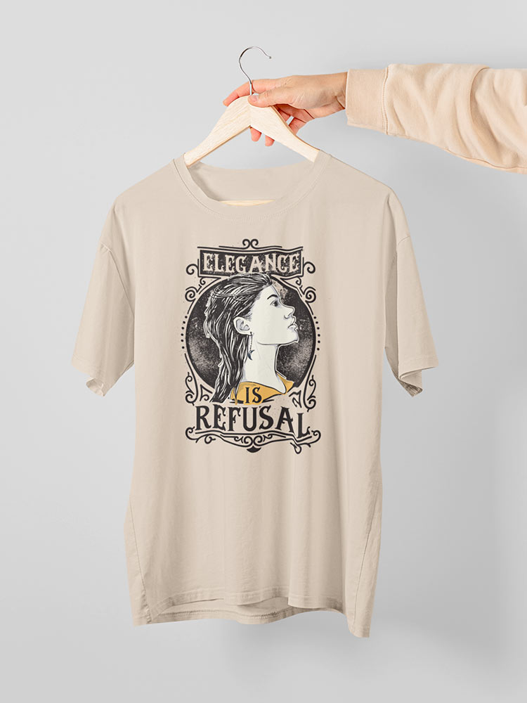 A person's hand holds a beige T-shirt with a graphic saying "Elegance is Refusal" surrounding a profile of a person, on a hanger against a gray background.