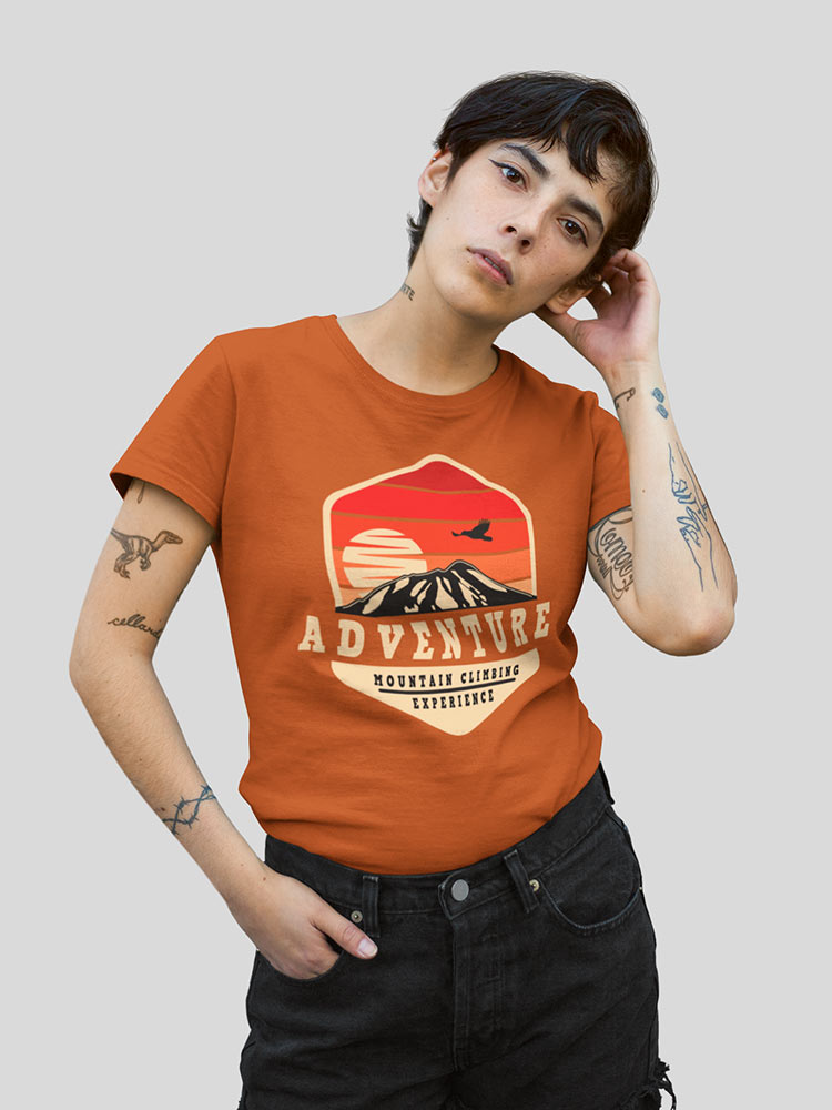 A person with short hair and tattoos wears an orange "Adventure" T-shirt, posing with hand on hip and touching their face against a grey background.