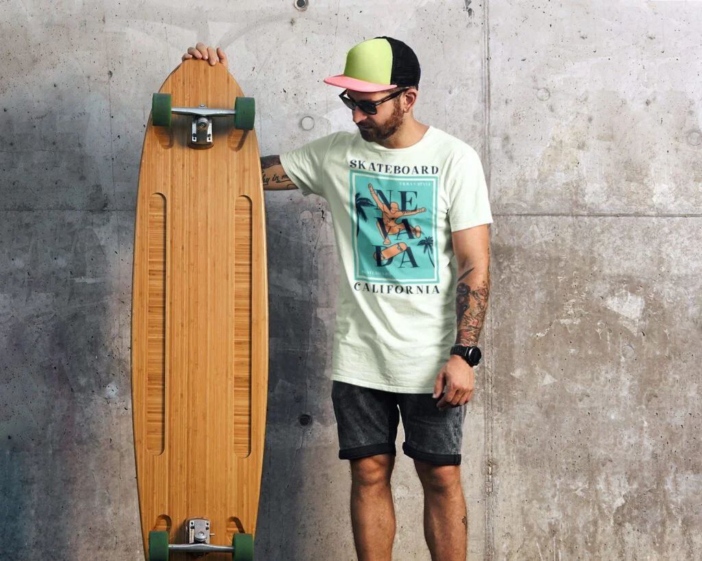 A person with tattoos holds a longboard, wearing a cap, sunglasses, and a t-shirt with "California" text. Concrete wall background suggests urban setting.