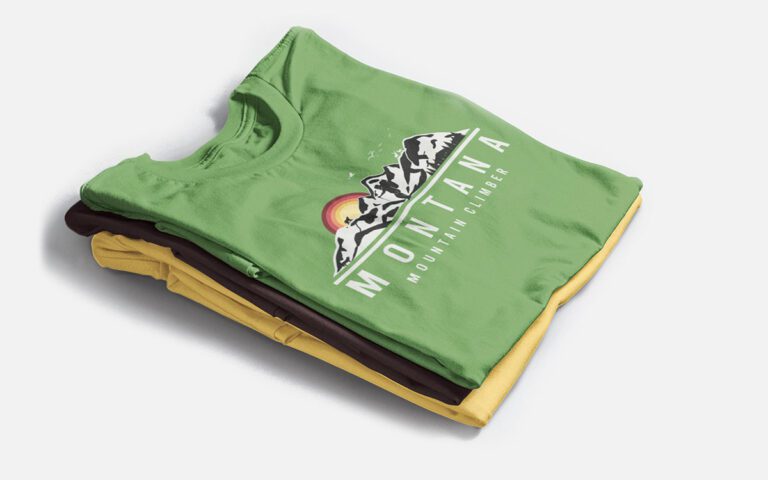 A neatly folded stack of three t-shirts in green, yellow, and brown, with the top green shirt displaying a mountain-themed graphic design.